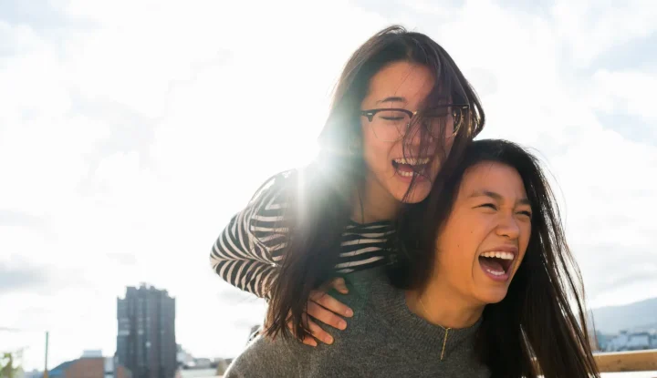 two women joyfully laughing and smiling together on a rooftop, enjoying a sunny day and each other's company.